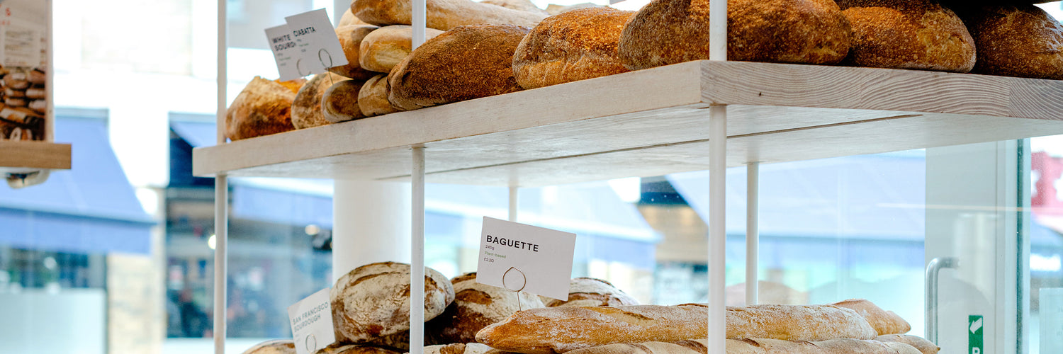 A display of various freshly baked loaves of bread and baguettes on shelves inside a bright, modern bakery with large windows.